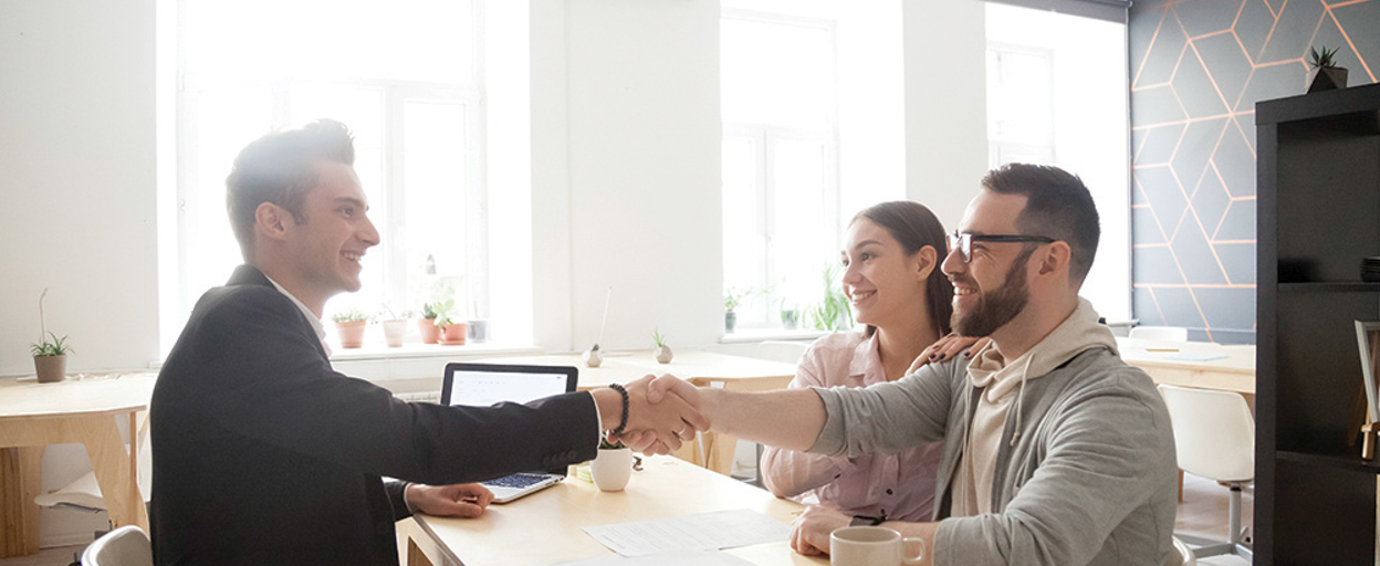 Couple smiling whilst man shakes hand with agent at desk