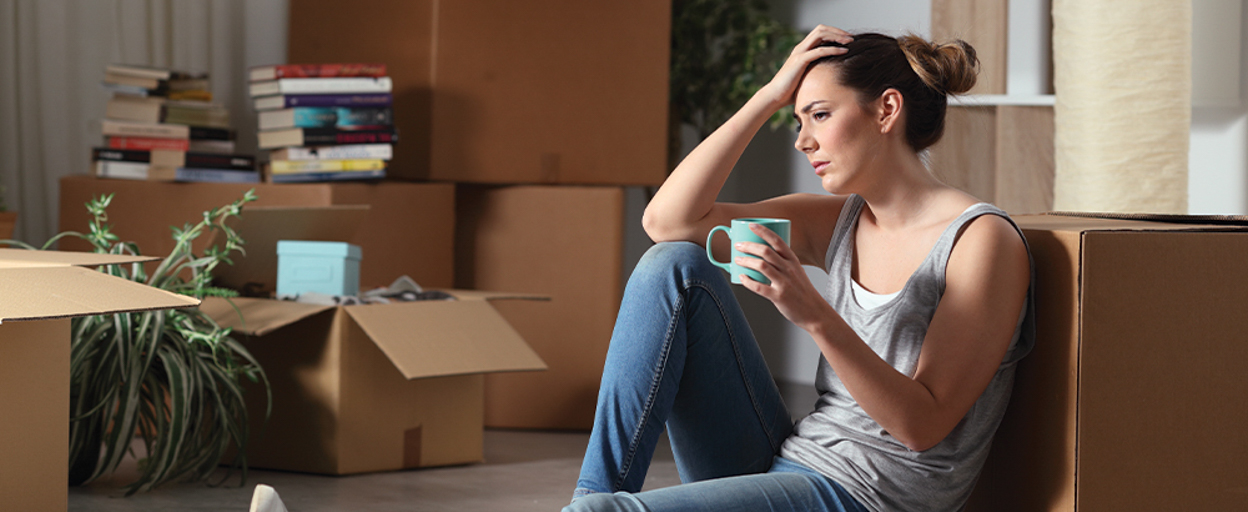 Woman sitting against moving box with coffee and hand against head
