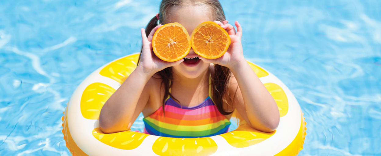 Girl in yellow floaty in pool holding oranges over eyes