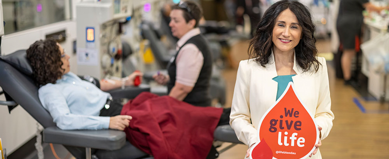 Antonia Mercorella standing in front of Laura Valenti donating blood, holding a sign that reads 'We Give Life'