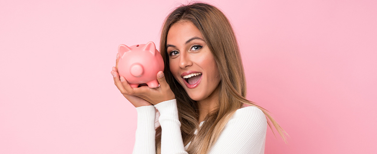 Female smiling while holding up a pink piggy bank with a pink background